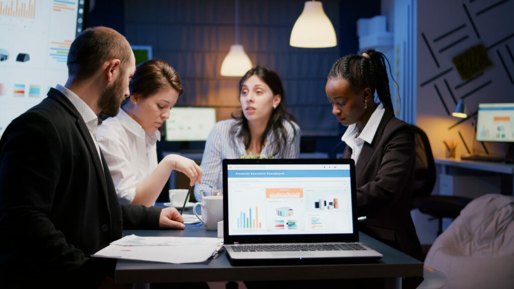 laptop computer with financial graphs presentation monitor standing conference table meeting room late night scaled.jpg